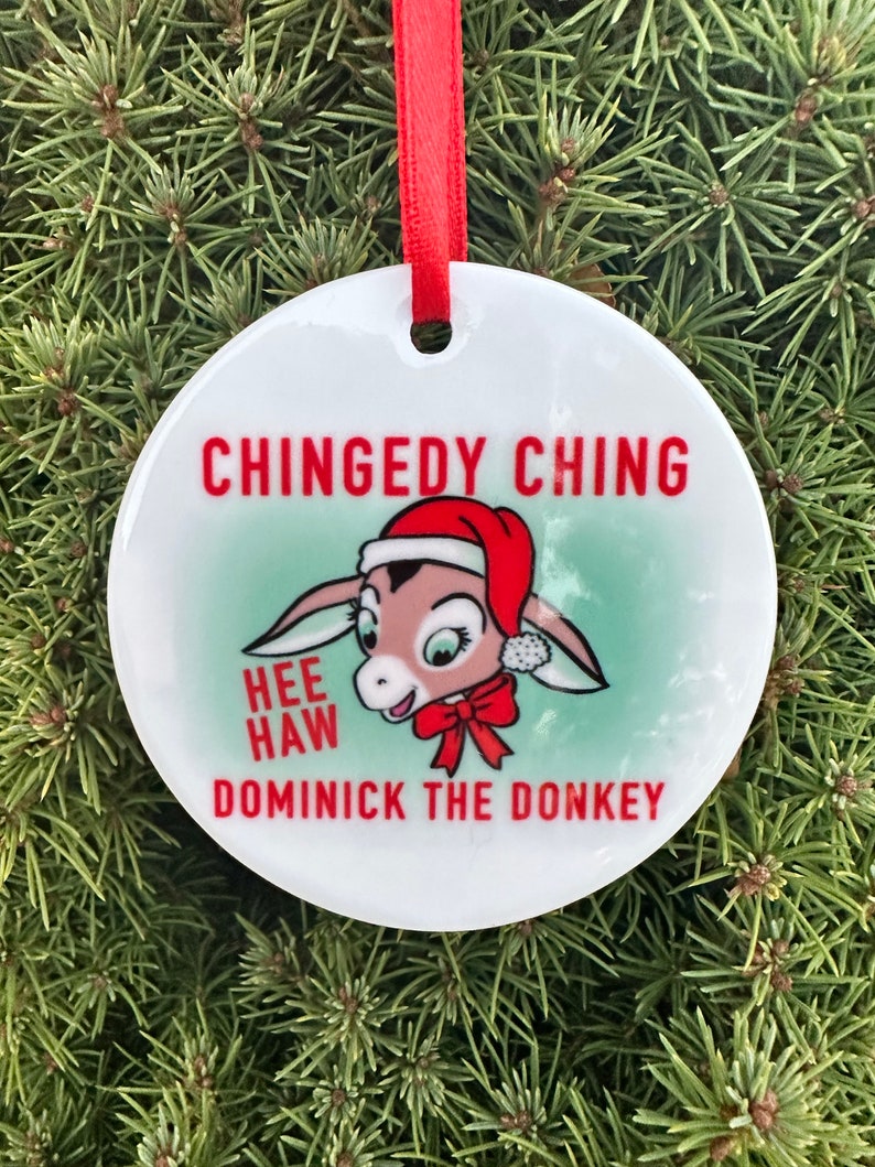 May include: A white round Christmas ornament with a red ribbon. The ornament features a cartoon donkey wearing a Santa hat and a red bow. The text on the ornament reads "Chingedy Ching Hee Haw Dominick the Donkey".