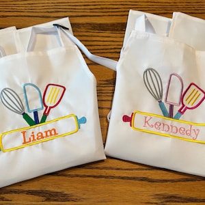 May include: Two white aprons with embroidered cooking utensils and personalized names. One apron says "Liam" in orange, and the other says "Kennedy" in pink. Each apron has a whisk, spatula, and scraper.