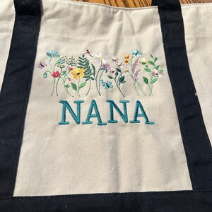 May include: A cream-colored canvas tote bag with black handles and a black base. The bag features an embroidered design of colorful wildflowers, butterflies, and a dragonfly above the teal-colored embroidered text that reads NANA.