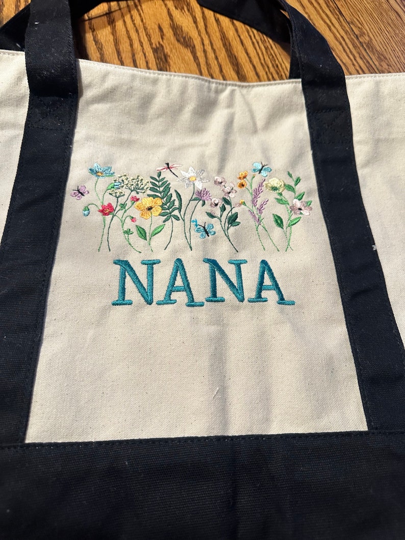 May include: A cream-colored canvas tote bag with black handles and a black base. The bag features an embroidered design of colorful wildflowers, butterflies, and a dragonfly above the teal-colored embroidered text that reads NANA.