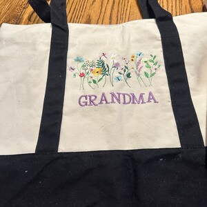 May include: A cream-colored canvas tote bag with black handles and a black base. The front features an embroidered design of colorful wildflowers, butterflies, and a dragonfly above the purple embroidered text that reads GRANDMA.