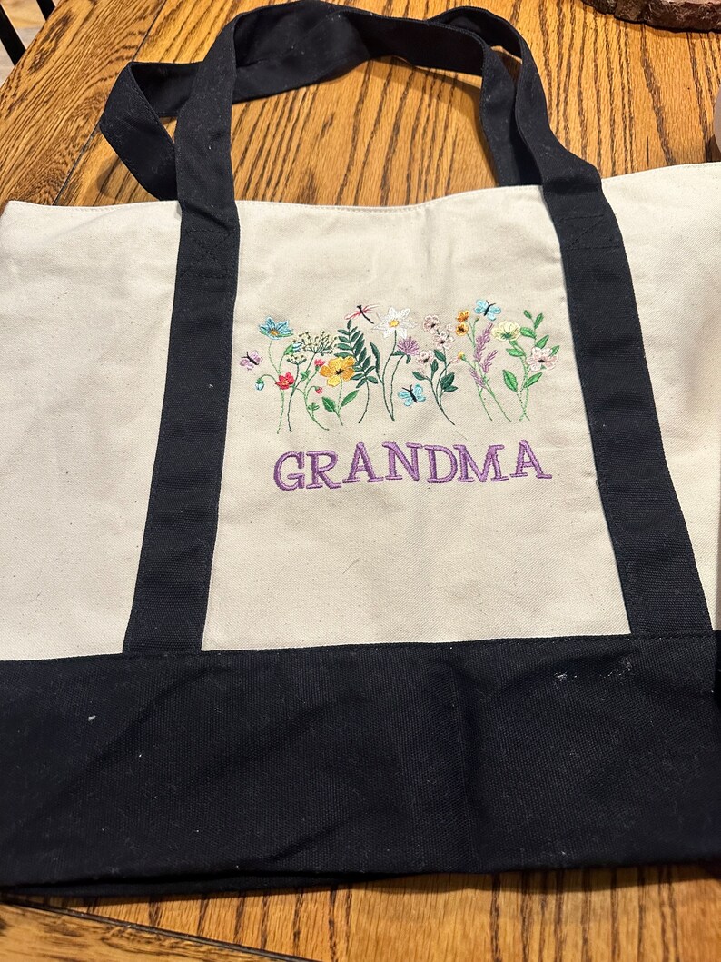 May include: A cream-colored canvas tote bag with black handles and a black base. The front features an embroidered design of colorful wildflowers, butterflies, and a dragonfly above the purple embroidered text that reads GRANDMA.
