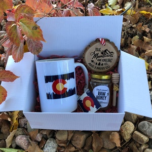 May include: A white gift box filled with a Colorado themed mug, a small jar of raw honey, a wooden ornament with the text "The Mountains Are Calling And I Must Go", and a Colorado sticker.
