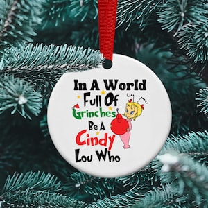 May include: White round Christmas ornament with the text "In a world full of Grinches be a Cindy Lou Who" and a cartoon image of Cindy Lou Who holding a red ornament.