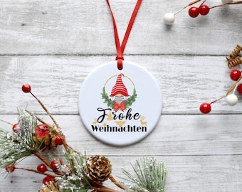 May include: White ceramic Christmas ornament with a red ribbon, featuring a gnome with a red hat and a wreath with gold accents. The ornament reads "Frohe Weihnachten" in black lettering.