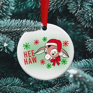 May include: A round Christmas ornament with a cartoon donkey wearing a Santa hat and a red bow. The ornament has the words "HEE HAW" in red and green snowflakes. A red ribbon is attached for hanging.