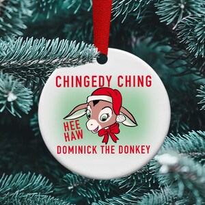 May include: A white round Christmas ornament with a cartoon donkey wearing a Santa hat and a red bow. The ornament has the text "Chingedy Ching Hee Haw Dominick the Donkey" printed on it.