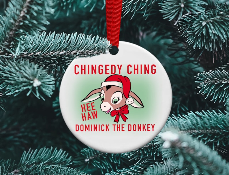 May include: A white round Christmas ornament with a cartoon donkey wearing a Santa hat and a red bow. The ornament has the text "Chingedy Ching Hee Haw Dominick the Donkey" printed on it.