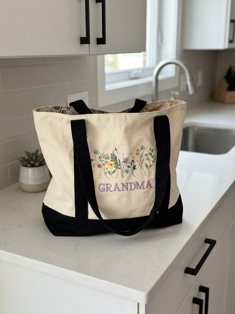 May include: A cream-colored canvas tote bag with black handles and a black base. The front features an embroidered design of colorful wildflowers and butterflies above the purple embroidered text that reads GRANDMA in capital letters.