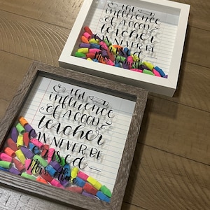 May include: Two square shadow box frames, one white and one dark wood, each containing a collection of colorful pencil erasers at the bottom. The glass front features black text reading, "the influence of a good teacher can never be erased" with names.