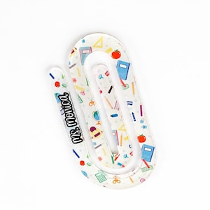 May include: A clear acrylic paperclip-shaped bookmark featuring a colorful pattern of school supplies including apples, books, pencils, and calculators. The text "ms. monica" is printed in black script on the left side of the bookmark.