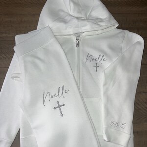 Puede incluir: Conjunto de sudadera con capucha y pantal&oacute;n de ch&aacute;ndal blanco con el nombre "Noelle" y una cruz bordada en la parte delantera. El pantal&oacute;n de ch&aacute;ndal tiene el nombre "Noelle" y una cruz bordada en la pierna izquierda. La sudadera tiene la fecha "5-3-25" bordada en la manga izquierda.