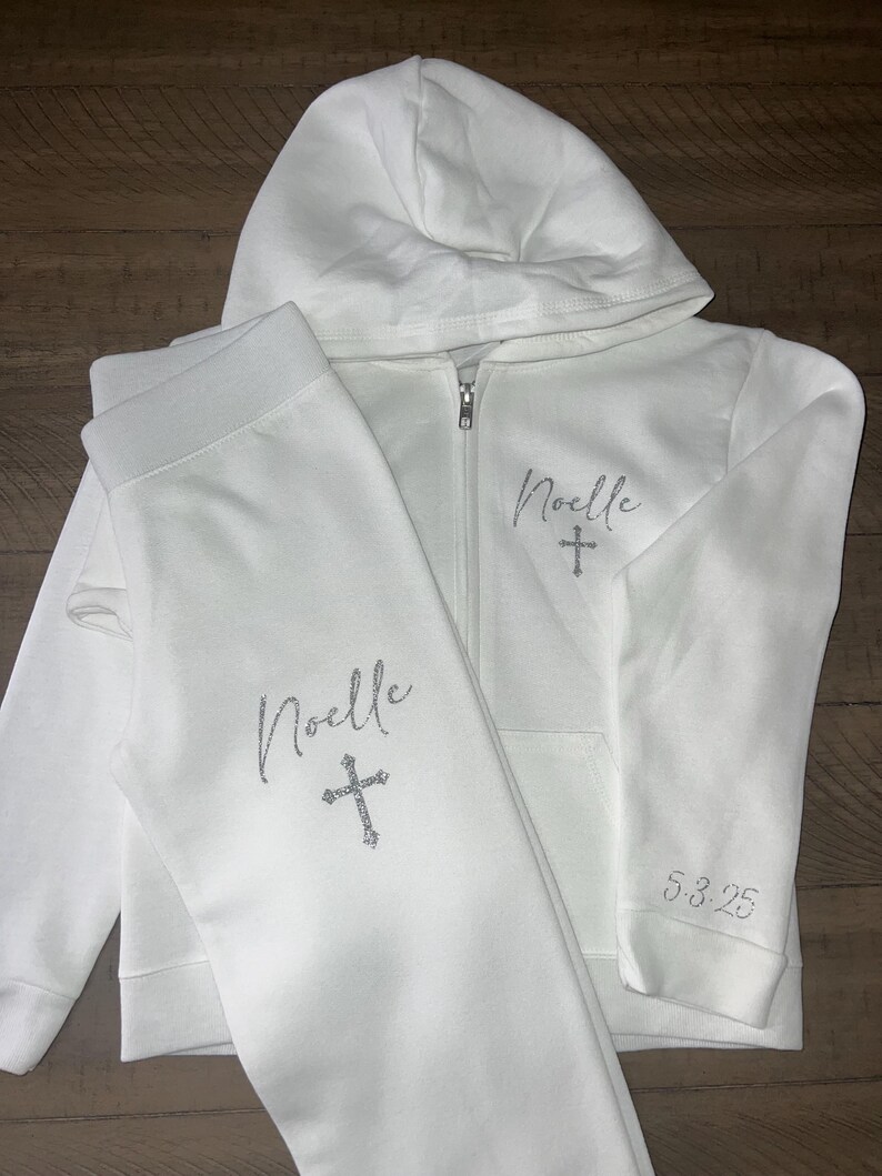 Puede incluir: Conjunto de sudadera con capucha y pantal&oacute;n de ch&aacute;ndal blanco con el nombre "Noelle" y una cruz bordada en la parte delantera. El pantal&oacute;n de ch&aacute;ndal tiene el nombre "Noelle" y una cruz bordada en la pierna izquierda. La sudadera tiene la fecha "5-3-25" bordada en la manga izquierda.
