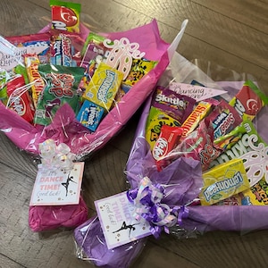 May include: Two candy bouquets wrapped in pink and purple tissue paper. Each bouquet contains various candy bars and treats. The bouquets are decorated with bows and tags that read "It's DANCE TIME! Good luck!"