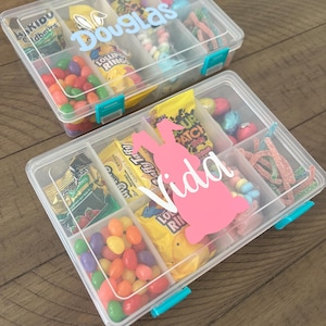 May include: Two clear plastic storage containers filled with various candies. The containers have blue lids and are labeled with the names "Douglas" and "Vida".