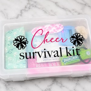 May include: A clear rectangular plastic storage box containing various small items, with the text "Cheer survival kit" printed on the lid in black and pink, flanked by two black pom-pom icons. The contents include a mint green patterned pouch, a pink floral nail file, a pack of Doublemint gum, and other small personal care items.