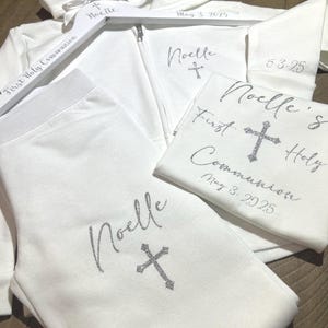 Puede incluir: Sudadera con capucha blanca, pantal&oacute;n y camiseta con el nombre "Noelle" y una cruz bordada en cada pieza. La camiseta tambi&eacute;n tiene el texto "Primera Comuni&oacute;n" y la fecha "3 de mayo de 2025".