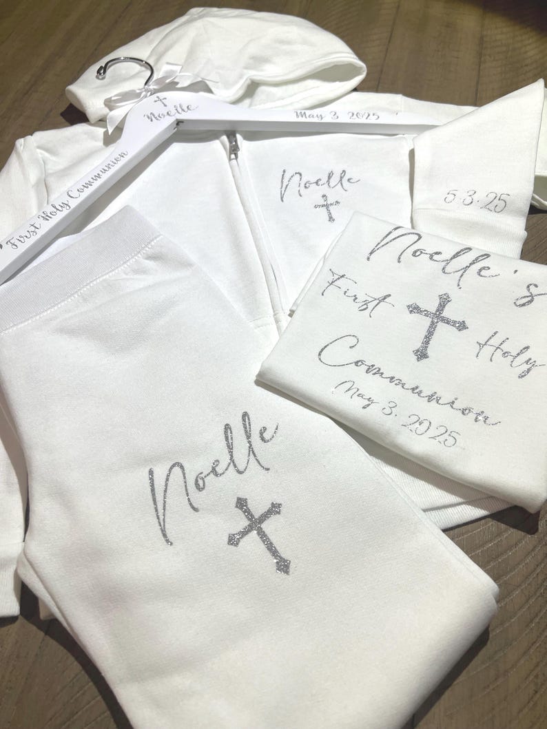 Puede incluir: Sudadera con capucha blanca, pantal&oacute;n y camiseta con el nombre "Noelle" y una cruz bordada en cada pieza. La camiseta tambi&eacute;n tiene el texto "Primera Comuni&oacute;n" y la fecha "3 de mayo de 2025".