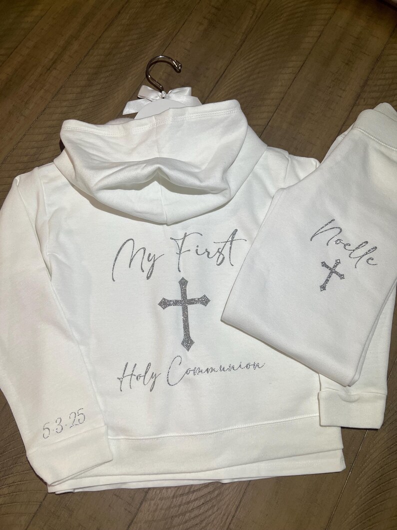 Puede incluir: Conjunto de sudadera con capucha y pantal&oacute;n de ch&aacute;ndal blanco con letras de purpurina plateada. La sudadera dice "My First Holy Communion" con una cruz y la fecha "5.3.25". El pantal&oacute;n de ch&aacute;ndal dice "Noelle" con una cruz.