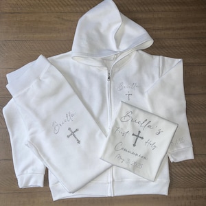 May include: White hooded sweatshirt and sweatpants set with a silver glitter design. The sweatshirt has a zipper closure and the sweatpants have an elastic waistband. The design includes the name "Briella's" and a cross with the text "First Holy Communion May 3, 2025".