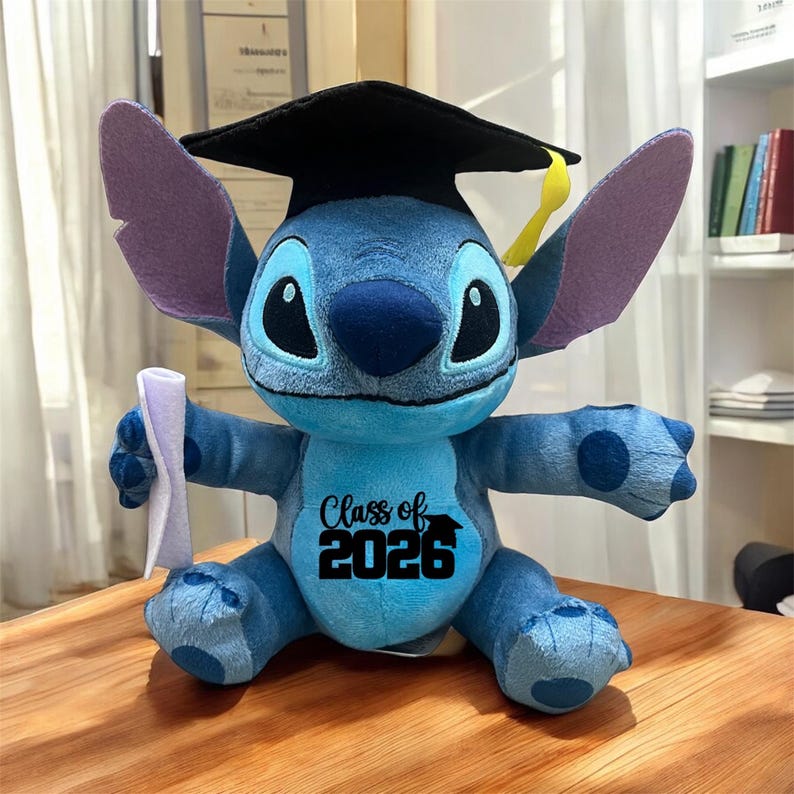 May include: A blue plush Stitch toy wearing a black graduation cap with a yellow tassel. The toy has large ears, big eyes, and holds a rolled-up diploma. The text "Class of 2026" is printed on its chest.