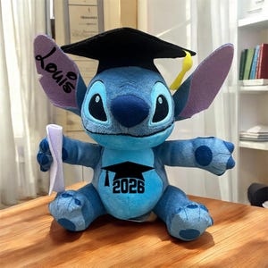 May include: A blue plush Stitch doll wearing a black graduation cap with a yellow tassel. The doll has the name "Louis" on one ear and the year "2026" with a graduation cap graphic on its chest. It holds a rolled-up diploma.