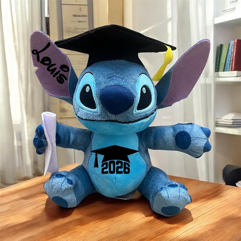 May include: A blue plush Stitch doll wearing a black graduation cap with a yellow tassel. The doll has the name "Louis" on one ear and the year "2026" with a graduation cap graphic on its chest. It holds a rolled-up diploma.
