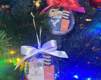 May include: Clear Christmas ornaments filled with miniature shopping bags and faux snow, adorned with pink and white satin bows. The ornaments hang on a decorated Christmas tree with colorful lights.