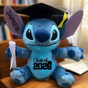 Puede incluir: Un peluche azul de Stitch con un birrete negro con borla amarilla. El peluche tiene el nombre "JAMES" en una oreja y "Class of 2026" impreso en el pecho. Sostiene un diploma enrollado.