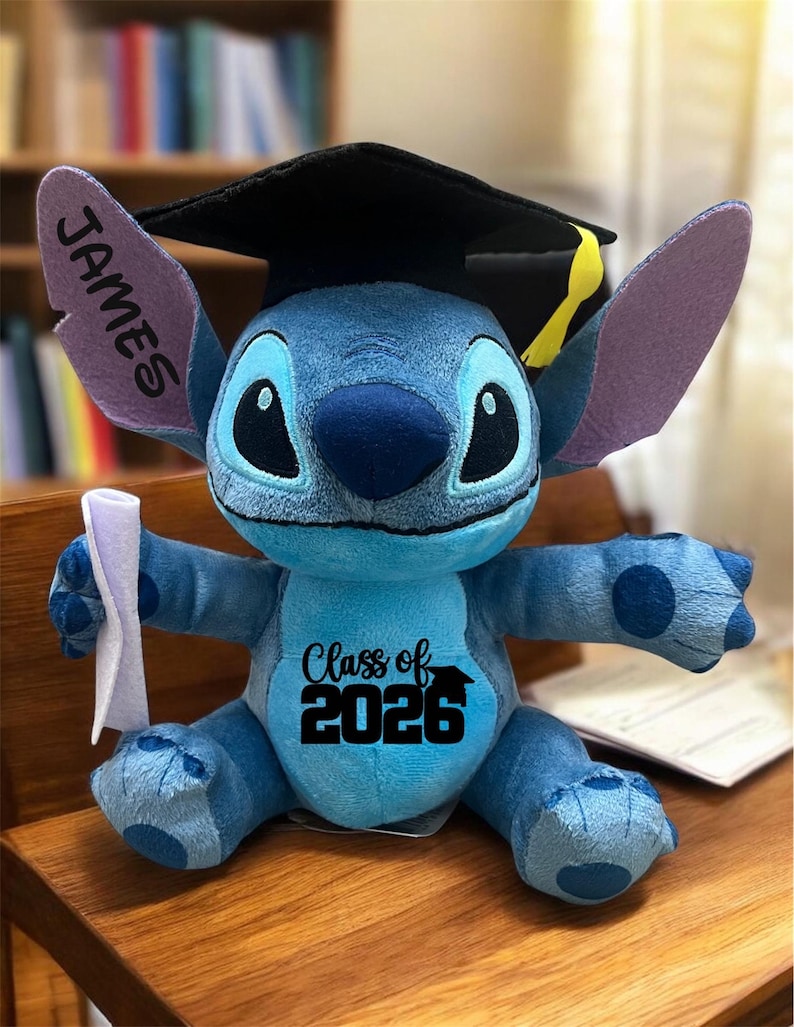 Puede incluir: Un peluche azul de Stitch con un birrete negro con borla amarilla. El peluche tiene el nombre "JAMES" en una oreja y "Class of 2026" impreso en el pecho. Sostiene un diploma enrollado.