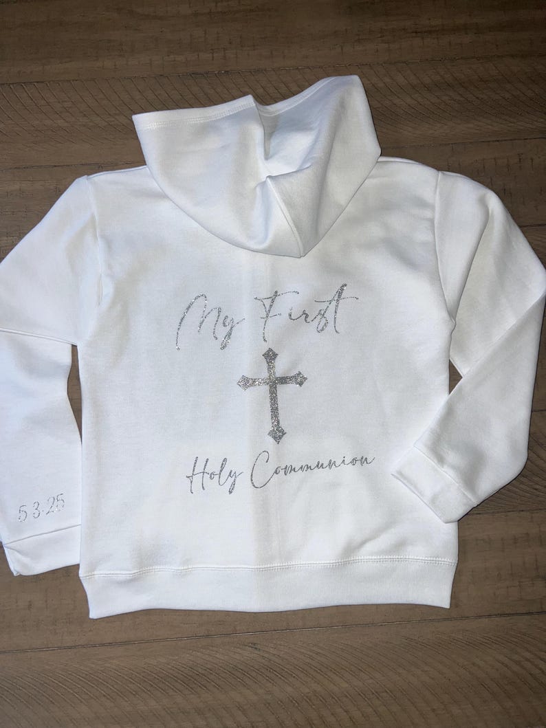 Puede incluir: Sudadera con capucha blanca con el texto "My First Holy Communion" y una cruz plateada con purpurina en la espalda.