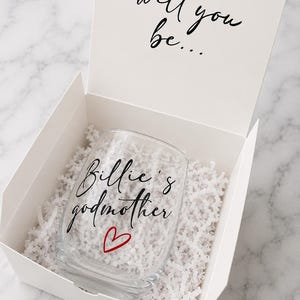 May include: A clear glass stemless wine glass nestled in a white gift box with white paper shred filler. The glass features black cursive text reading Billie's godmother and a small red heart. The box lid has black text reading will you be...