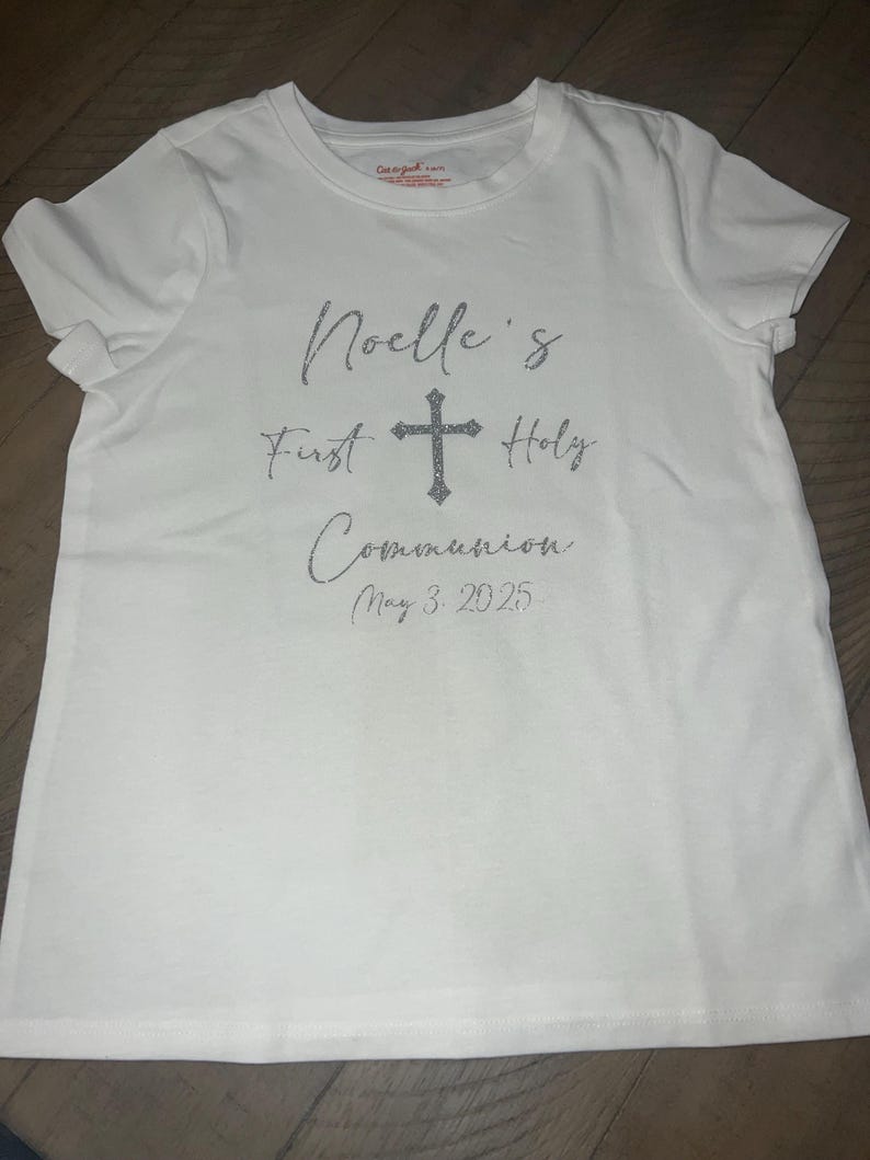 Puede incluir: Camiseta blanca con el texto "Noelle's First Holy Communion May 3, 2025" y una cruz de purpurina plateada.