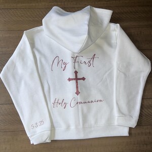 Puede incluir: Sudadera con capucha blanca con el texto "My First Holy Communion" y una cruz en purpurina rosa.