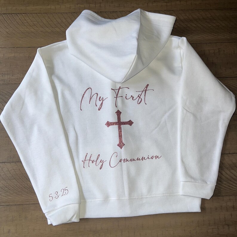 Puede incluir: Sudadera con capucha blanca con el texto "My First Holy Communion" y una cruz en purpurina rosa.