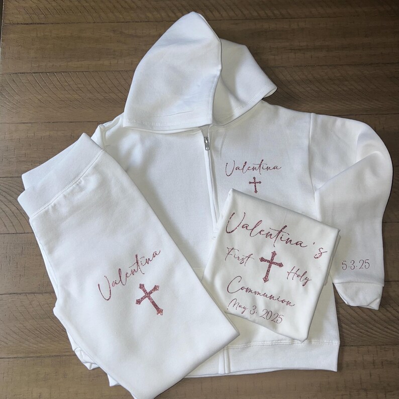 Puede incluir: Sudadera con capucha blanca, pantal&oacute;n de ch&aacute;ndal y camiseta con el nombre "Valentina" y una cruz bordada en purpurina rosa. La camiseta tambi&eacute;n dice "Primera Comuni&oacute;n" y la fecha "3 de mayo de 2025".