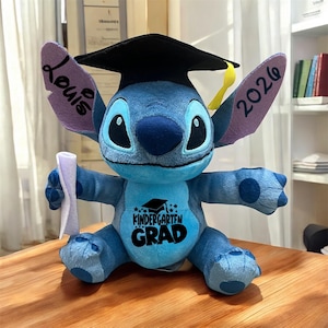May include: A blue plush Stitch toy wearing a black graduation cap and holding a diploma. The toy has the text "Kindergarten Grad" on its chest and "Louis 2026" on its ears.