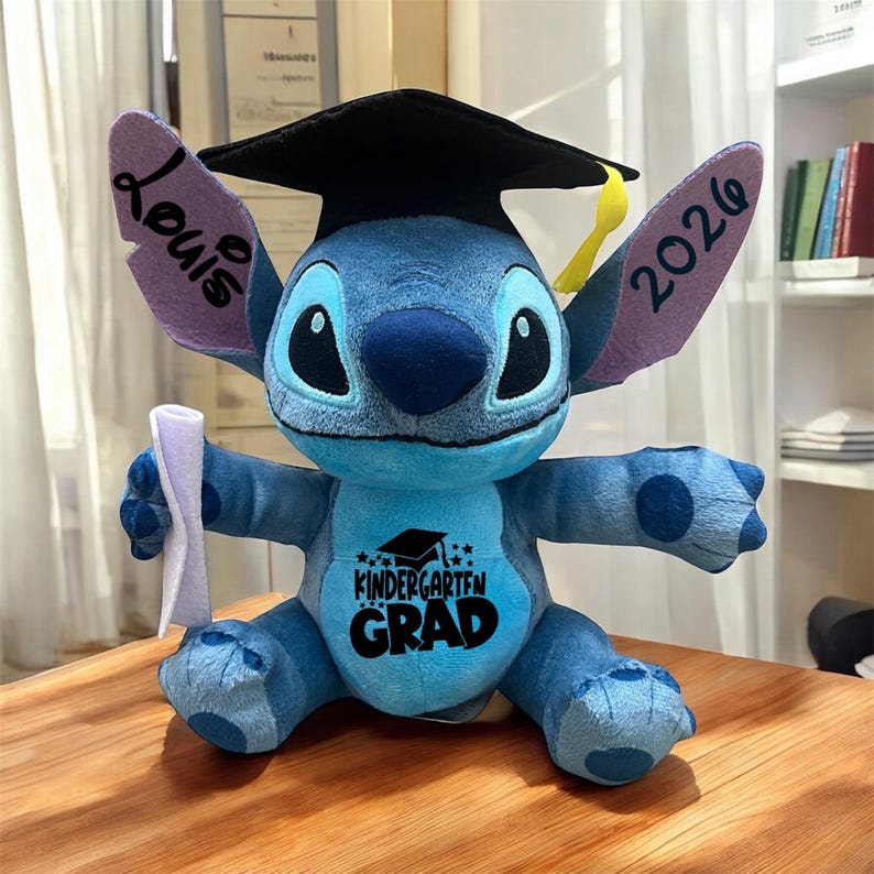 May include: A blue plush Stitch toy wearing a black graduation cap and holding a diploma. The toy has the text "Kindergarten Grad" on its chest and "Louis 2026" on its ears.