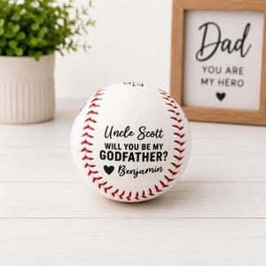 May include: A white baseball with red stitching and the text "Uncle Scott, will you be my Godfather?" in black. A small black heart and the name "Benjamin" are below the text. A framed sign that says "Dad, you are my hero" is in the background.