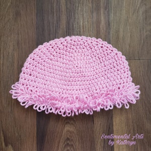 May include: A light pink crocheted hat with a ruffled edge. The hat is made of a soft yarn and has a rounded crown. The bottom edge features a decorative, looped trim. The hat is displayed on a wooden surface.