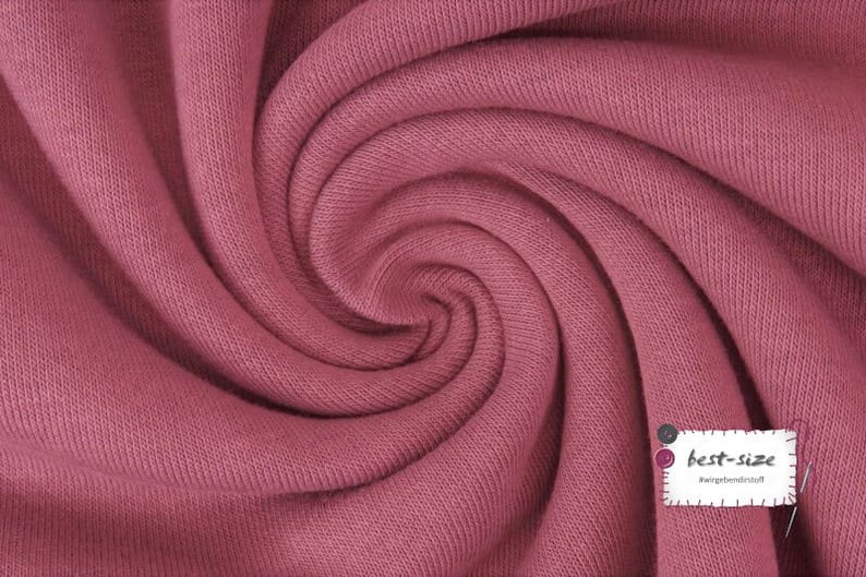 May include: Close-up of a soft, pink knitted fabric with a textured surface. The fabric is folded and twisted, creating a swirling pattern. A small white label with the text "best-size" is visible in the lower right corner.