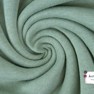 May include: Close-up of a soft, sage green fabric with a textured knit pattern. The fabric is folded and twisted, creating a swirling effect.