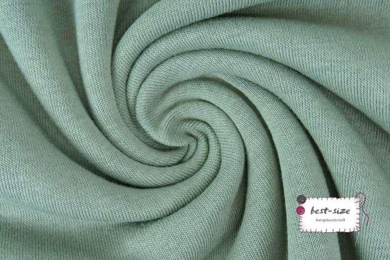 May include: Close-up of a soft, sage green fabric with a textured knit pattern. The fabric is folded and twisted, creating a swirling effect.