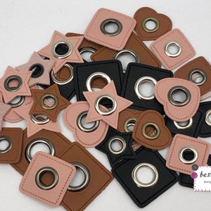 Könnte beinhalten: Sortiment aus Lederösen in Rosa, Braun und Schwarz mit silbernen und messingfarbenen Ösen. Die Ösen haben verschiedene Formen, darunter Sterne, Herzen und Quadrate.