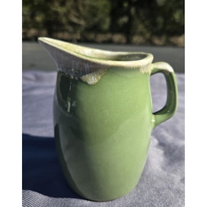 May include: A sage green ceramic pitcher with a smooth, glossy finish. The rim features a decorative drip-style glaze in a creamy off-white tone that extends slightly down the upper edge. It has a curved handle and a wide, molded pouring spout.