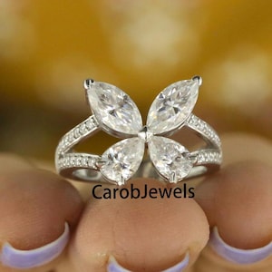 Pode incluir: Um anel de prata com um design de borboleta com cinco pedras claras em forma de marquise. O anel é adornado com pedras claras menores ao redor da banda. O nome da marca "CarobJewels" é visível no anel.