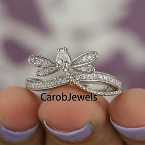 May include: A silver ring featuring a dragonfly design with clear cubic zirconia stones. The dragonfly's wings are spread out and the body is adorned with smaller stones. The ring band is thin and has a delicate design.