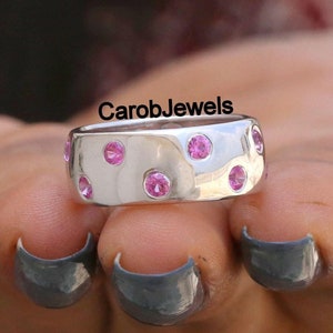 Puede incluir: Un anillo de banda de plata con un acabado martillado y piedras preciosas rosas engastadas alrededor de la banda. El texto "CarobJewels" es visible en el anillo.
