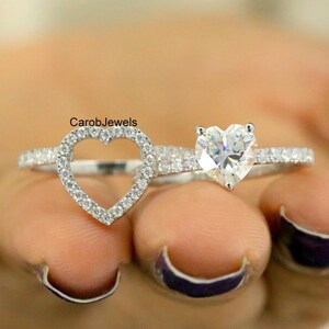 May include: A silver-toned ring set featuring two bands. One band has a heart-shaped outline encrusted with small clear stones, and the other band features a single heart-shaped clear stone. The text CarobJewels is visible above the rings.