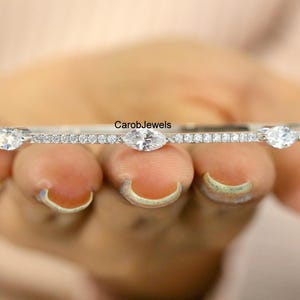 Op de afbeelding: Een zilverkleurige armband met drie ovale edelstenen en talrijke kleinere, ronde stenen. De armband wordt in een hand gehouden, met de merknaam "CarobJewels" zichtbaar.